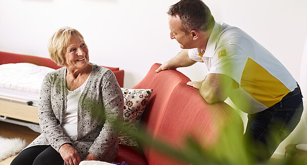 Hilfswerk-Mitarbeiter redet mit einer älteren Dame auf einem Sofa.